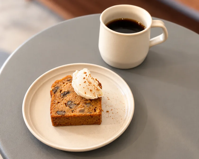 青灰色の丸テーブルに、クリームを添えたキャロットケーキの皿と、白いマグカップのコーヒーが並ぶ